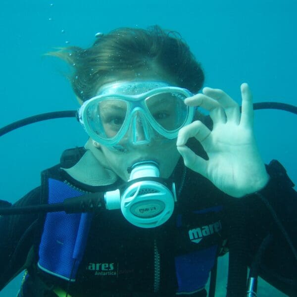 Child scuba diving underwater, wearing scuba gear and making an OK hand signal representing scuba diving for kids.