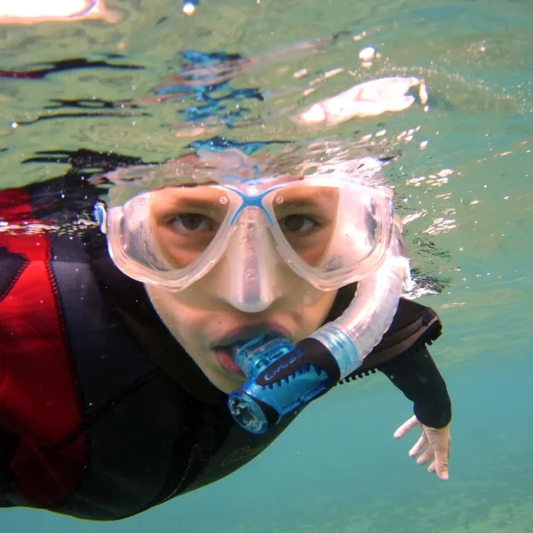 Person snorkeling underwater wearing a mask and blue snorkel, in a red and black wetsuit, facing the camera.